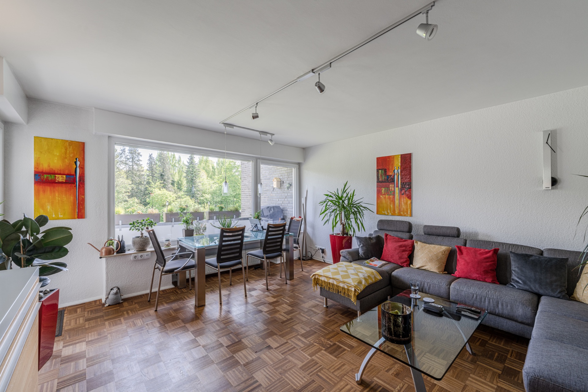 Helles Wohnzimmer mit Ecksofa, Glastisch, Essbereich, Pflanzen und großen Fenstern zum Garten.