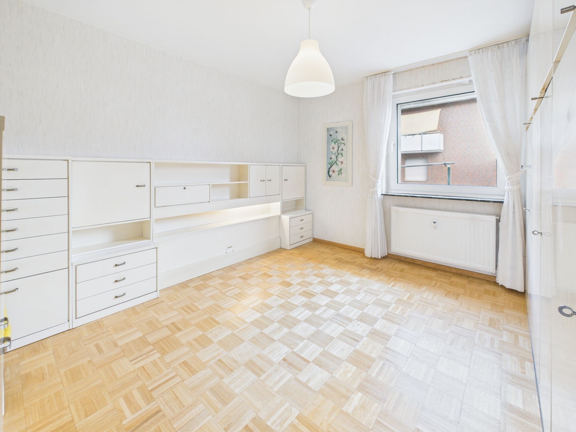 Helles Zimmer mit Parkettboden, weißen Einbauschränken und großem Fenster mit Vorhängen.