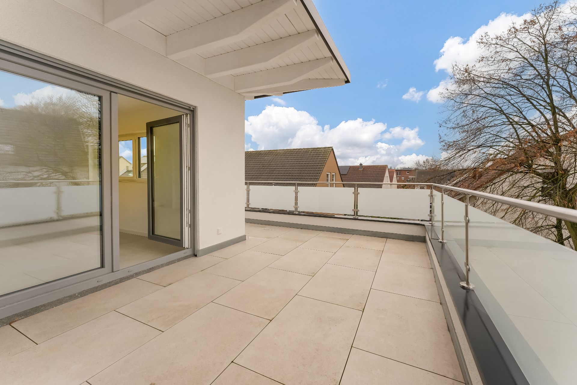 Große, geflieste Terrasse mit Glasgeländer und Blick auf Nachbarhäuser und Bäume.