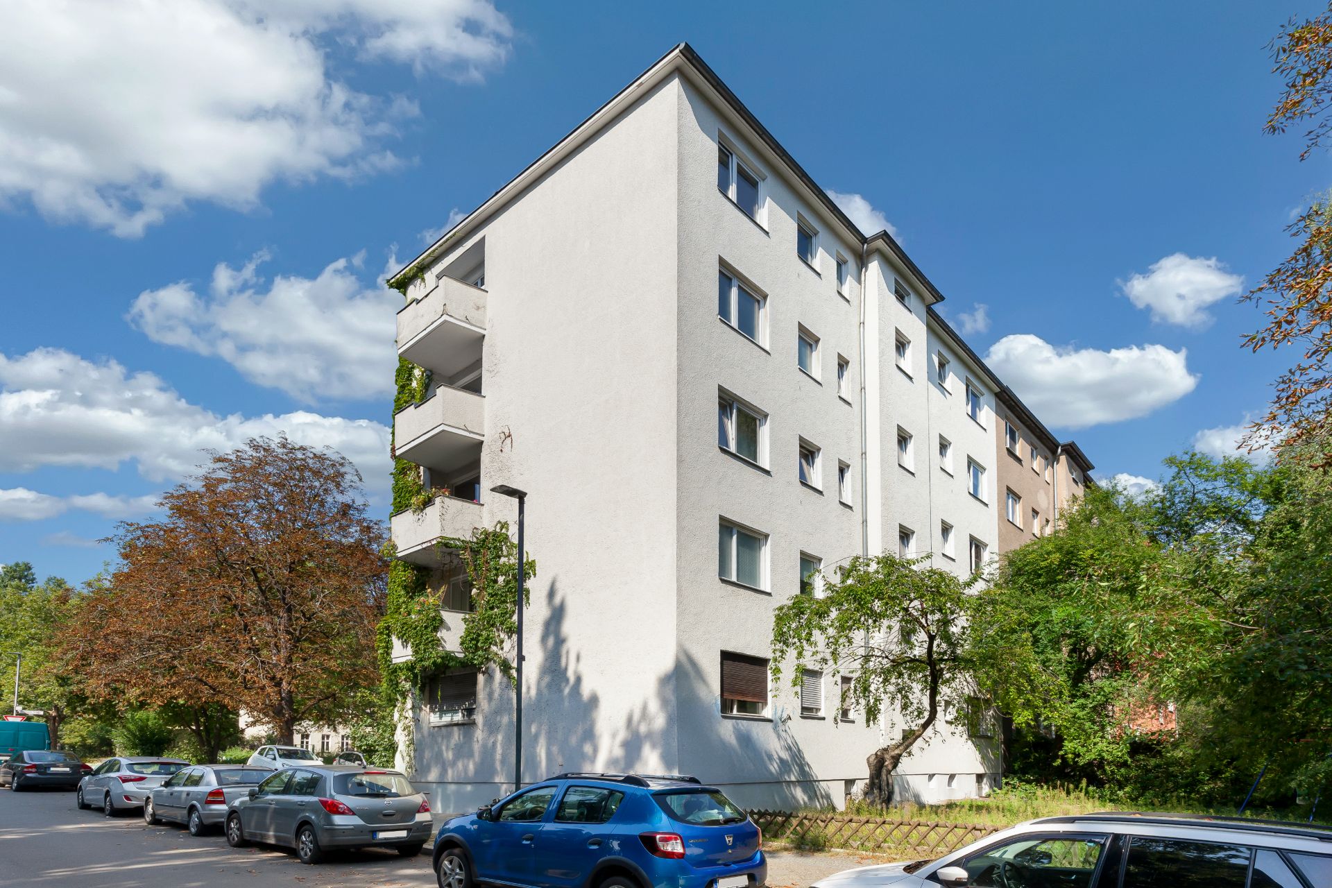 Mehrstöckiges Wohnhaus mit Balkonen, parkenden Autos und Bäumen an einer Straße.