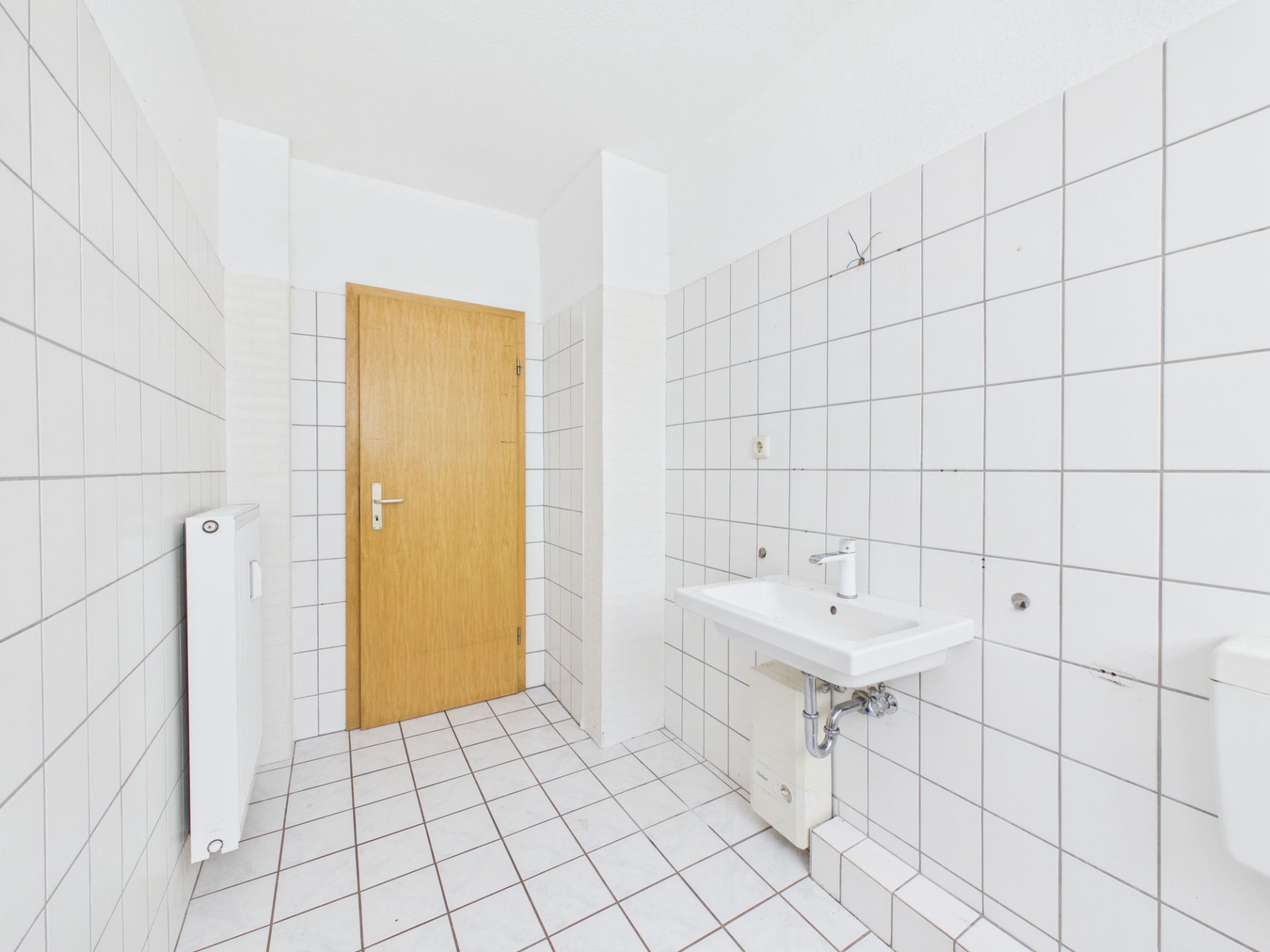 Weiß gefliestes Badezimmer mit Waschbecken, Heizung und Holztür.