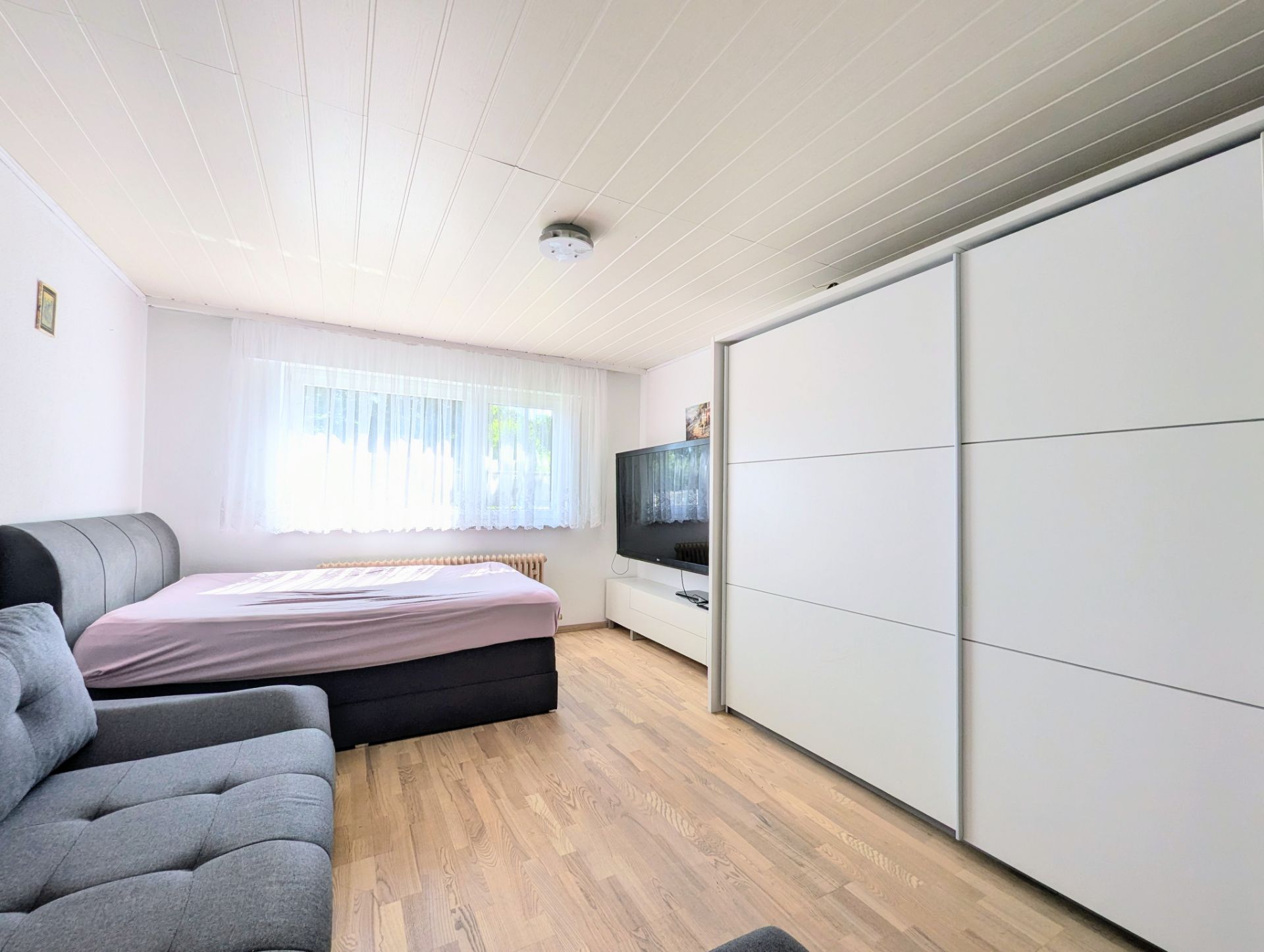 Helles Schlafzimmer mit Bett, Sofa, großem Schrank und Fernseher auf Holzfußboden.