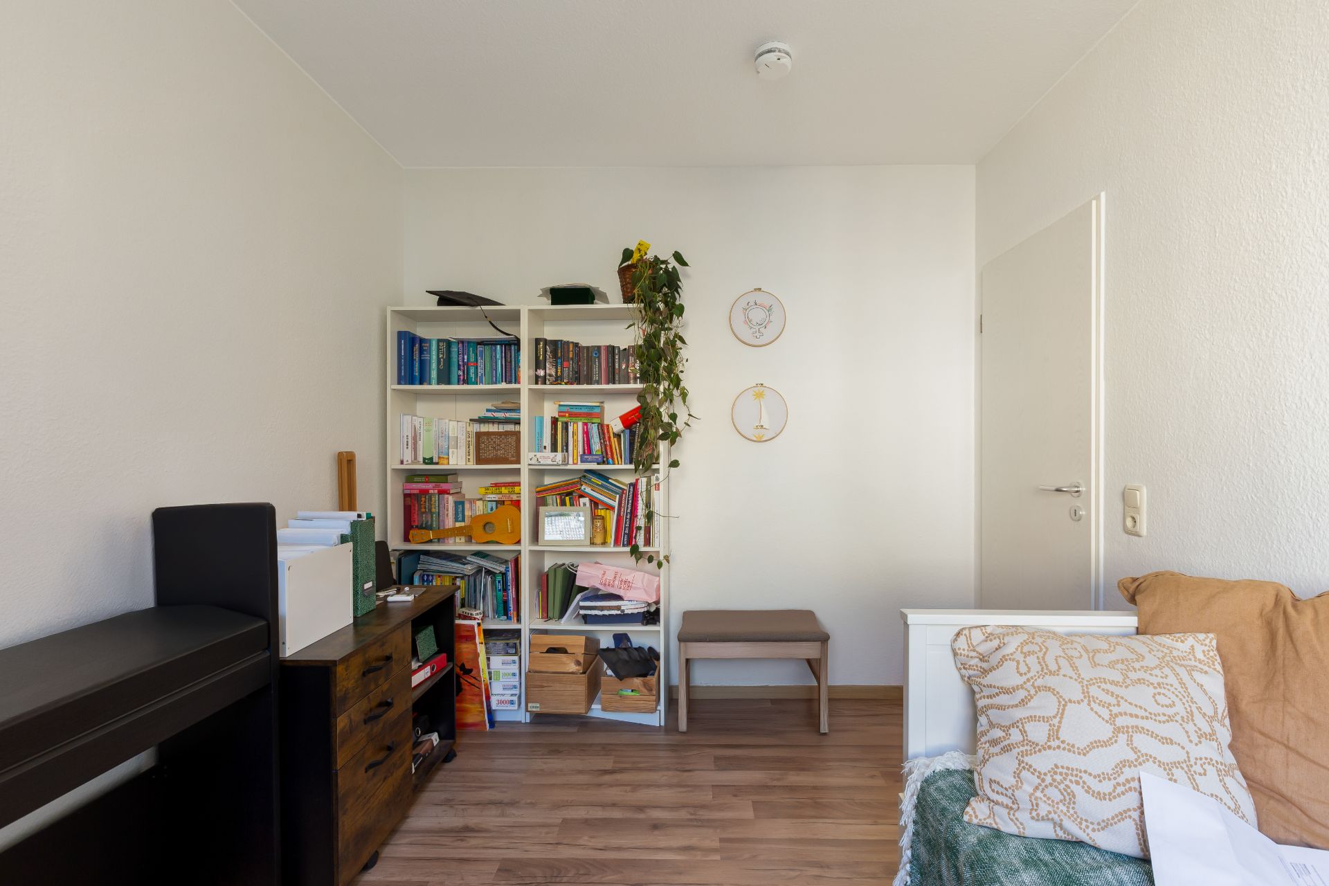 Helles Zimmer mit Bücherregal, kleiner Bank, Schreibtisch und Sofa mit Kissen.