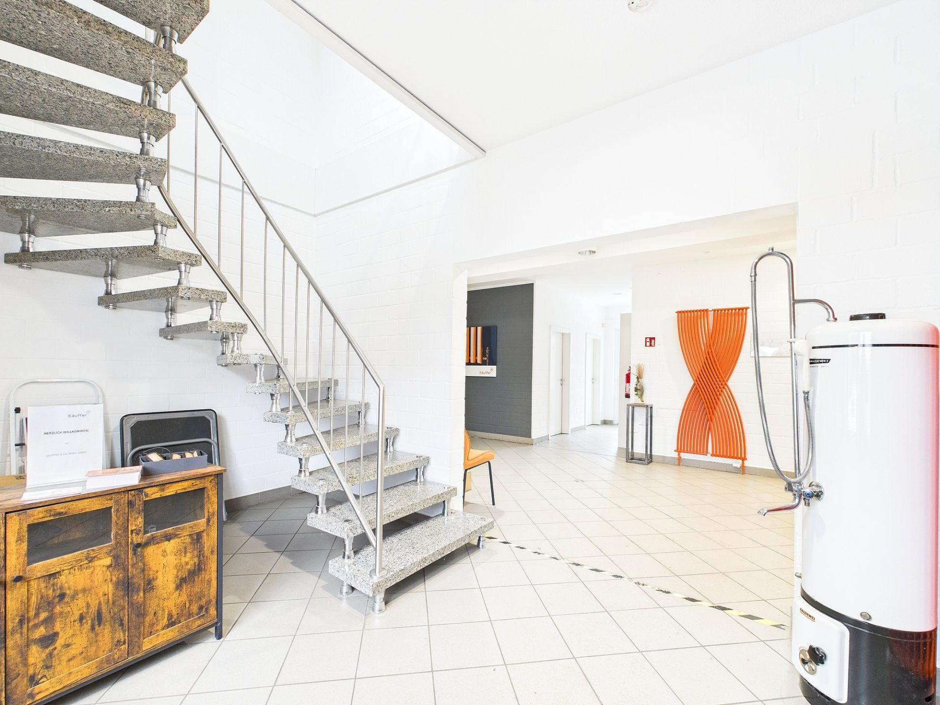 Heller Flur mit Treppe, Holzmöbel, Stuhl und großem Wasserboiler, Fliesenboden.