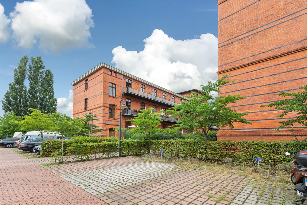 Backsteingebäude mit Balkonen, davor Parkplätze, Bäume und Büsche bei sonnigem Wetter.