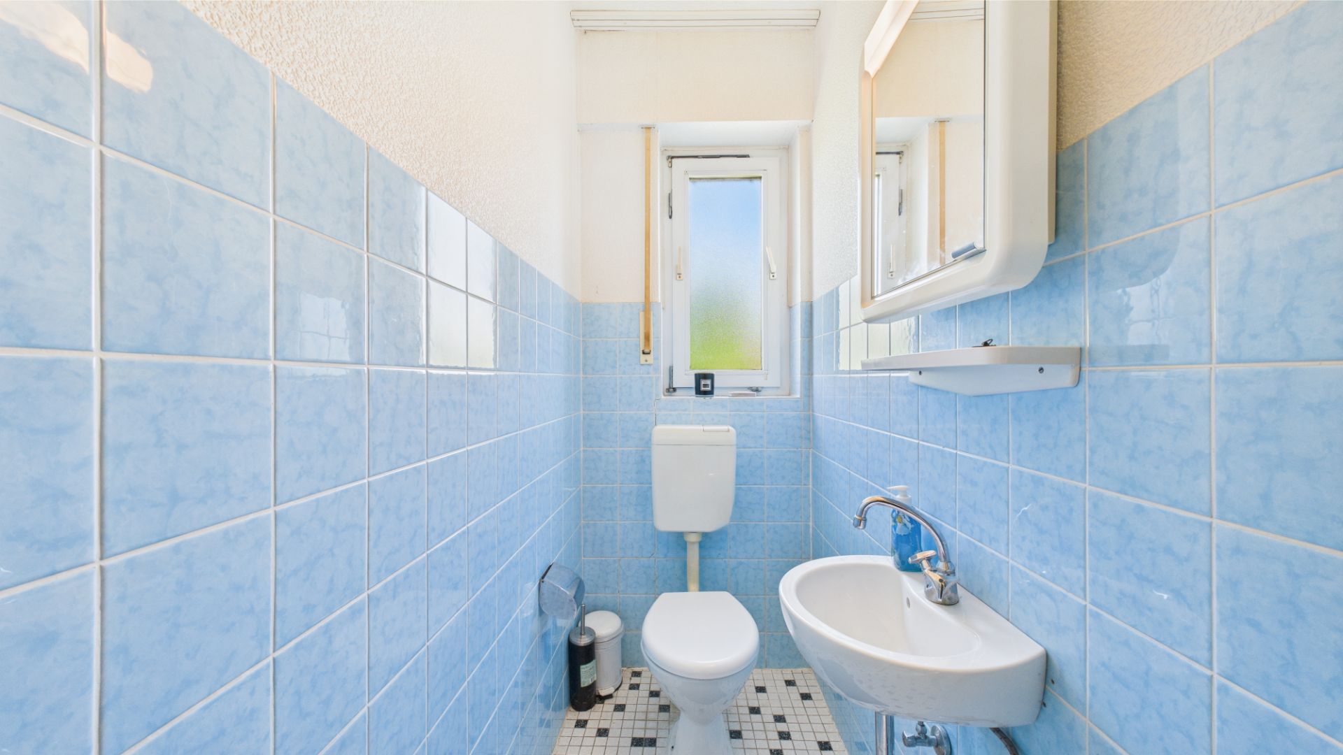 Schmales Badezimmer mit blauen Fliesen, WC, kleinem Waschbecken und Fenster.