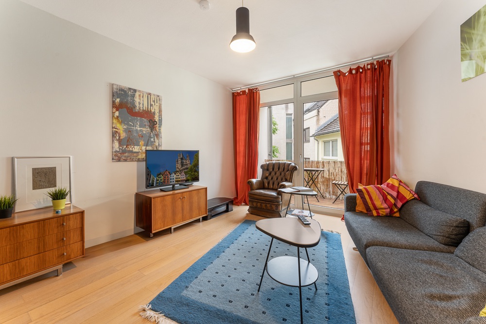 Modernes Wohnzimmer mit grauem Sofa, Sessel, Fernseher, blauem Teppich und roten Vorhängen.