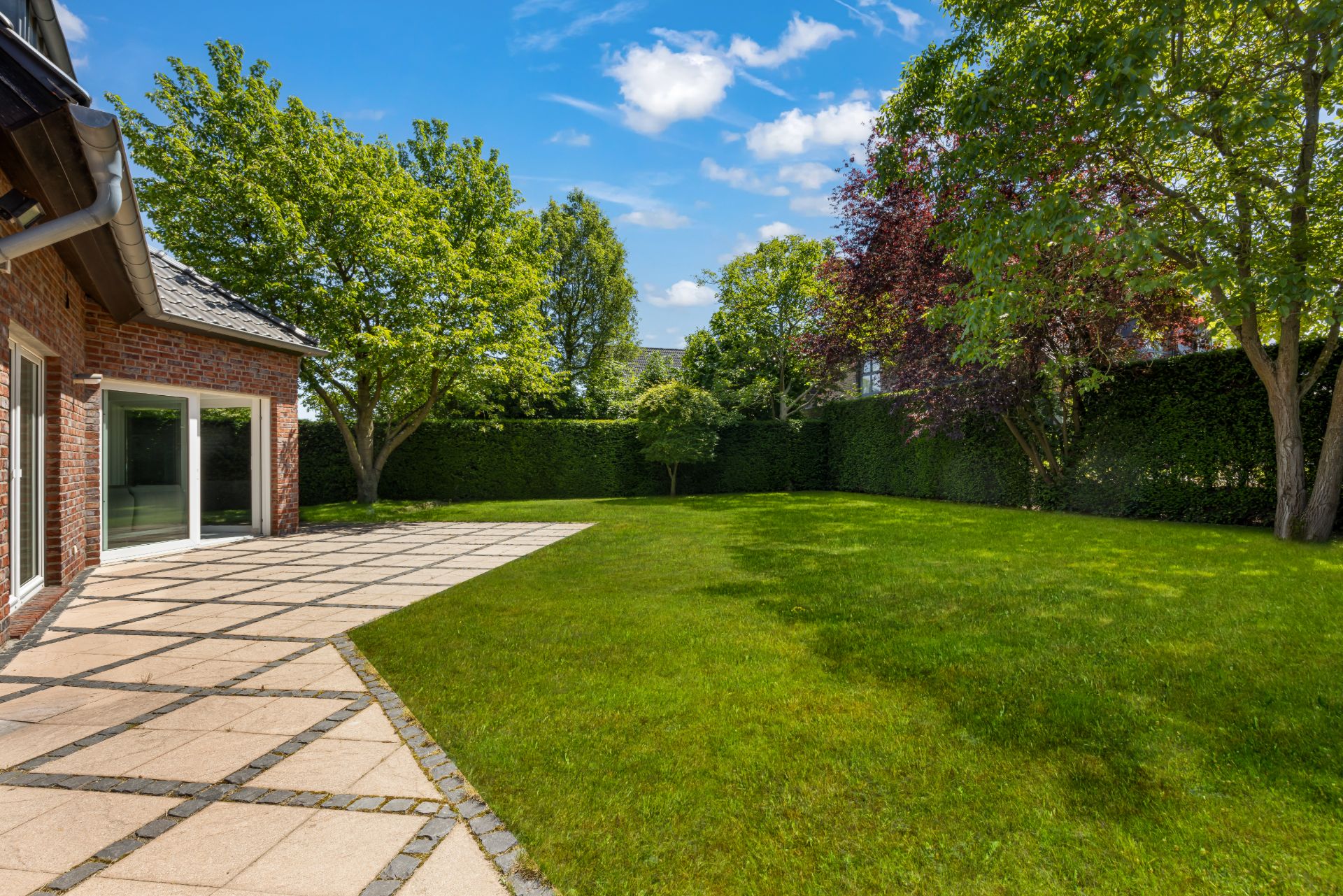 Grüner Garten mit Rasenfläche, Terrasse und mehreren Bäumen neben einem Haus.