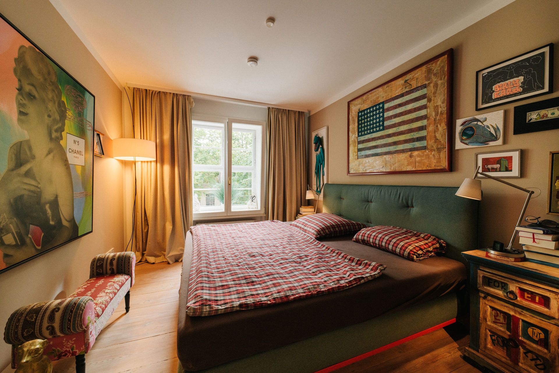 Schlafzimmer mit Doppelbett, großen Fenstern, Kunst an den Wänden und Nachttisch mit Lampe.
