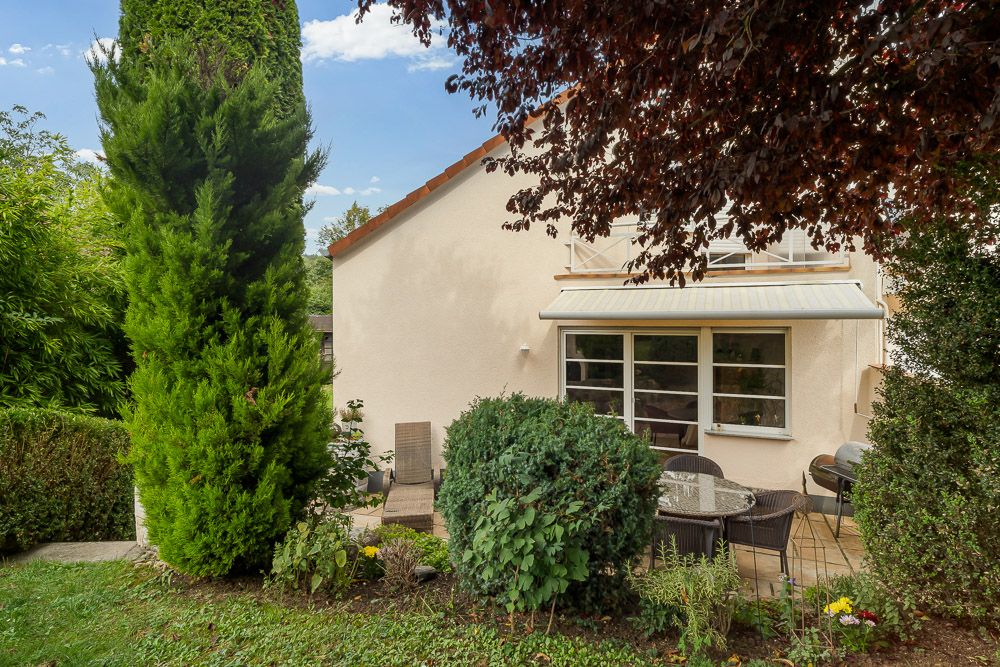Garten mit Terrasse, Esstisch und Stühlen vor einem hellen Wohnhaus mit großen Fenstern.