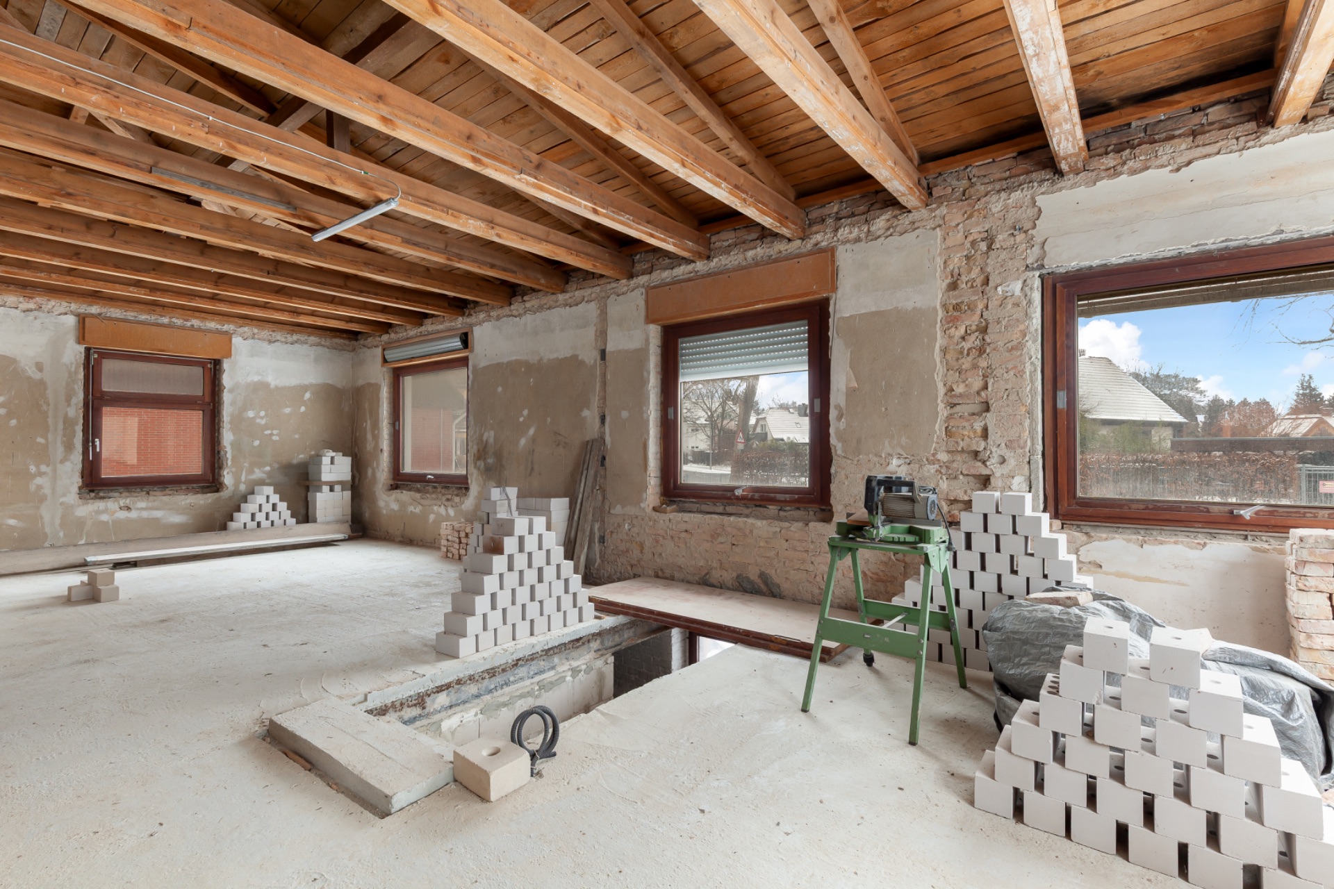 Großer, heller Raum im Rohbau mit sichtbaren Holzbalken und gestapelten Bauziegeln.