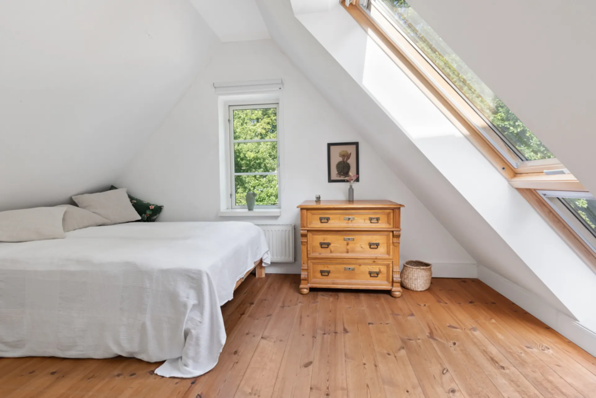 Helles Schlafzimmer mit Holzboden, Bett, Kommode und großen Dachfenstern.