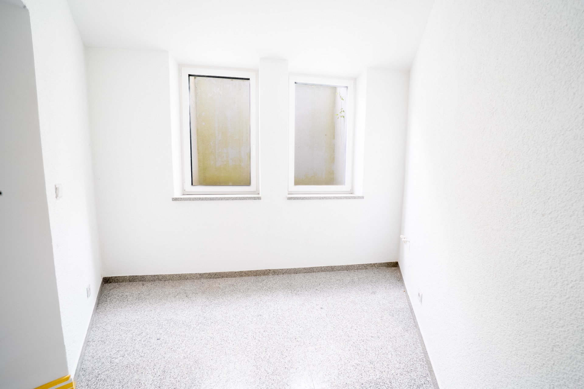 Kleiner leerer Raum mit zwei Fenstern und weiß gestrichenen Wänden.