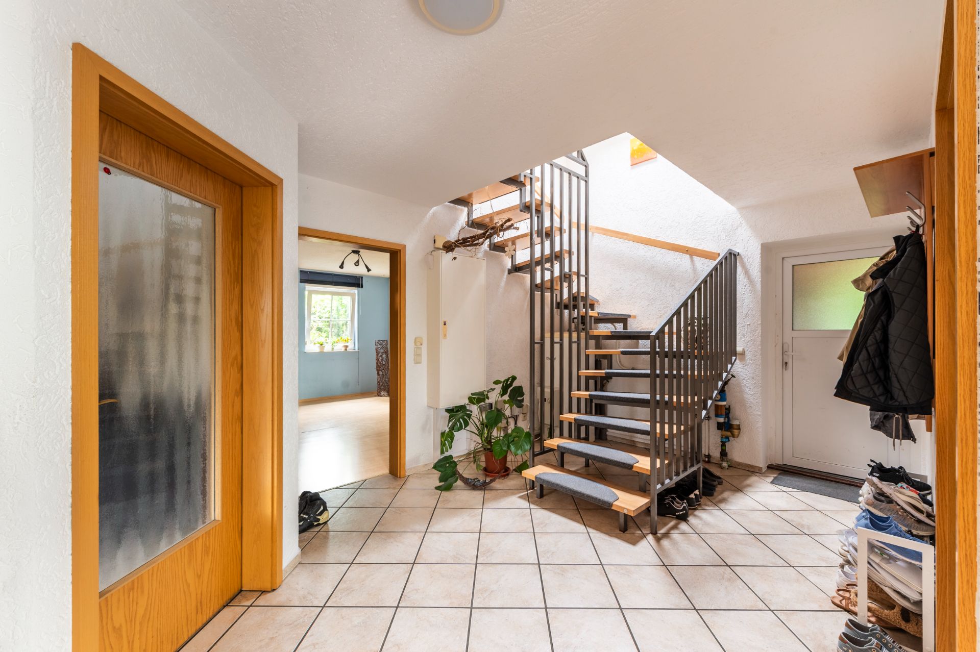 Hausflur mit moderner Holztreppe, offenen Türen, Fliesenboden und Pflanzen.