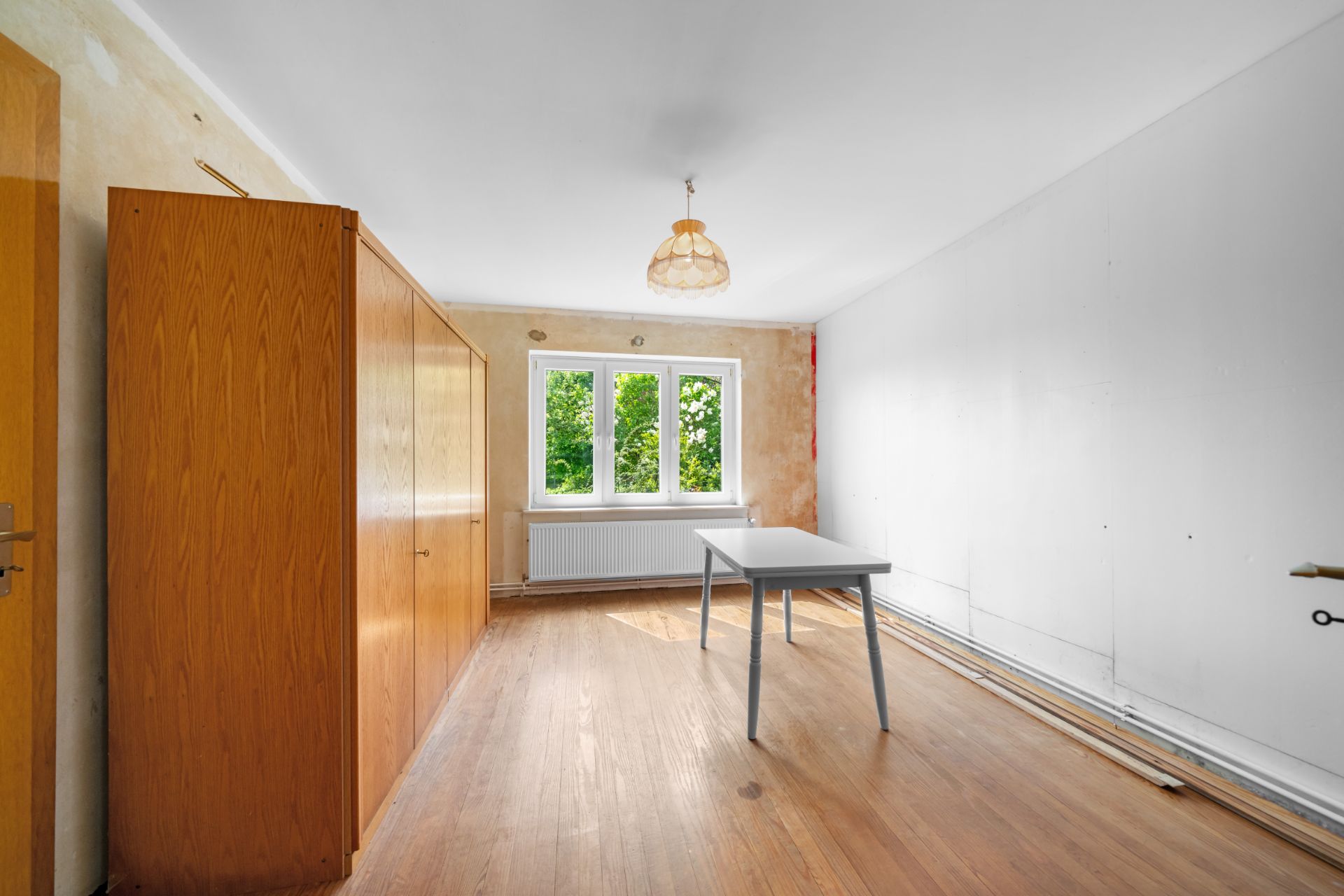 Ein leerer Raum mit Holzboden, kleinem Tisch, großem Schrank und Fenster, durch das Bäume sichtbar sind.