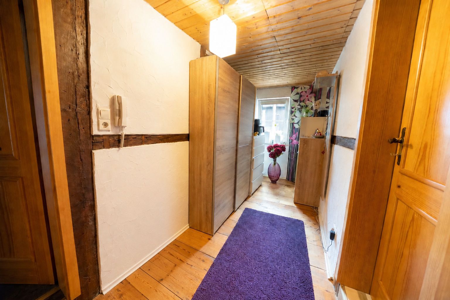 Heller Flur mit Holzboden, lila Teppich, Schränken und einer Vase mit Blumen am Fenster.