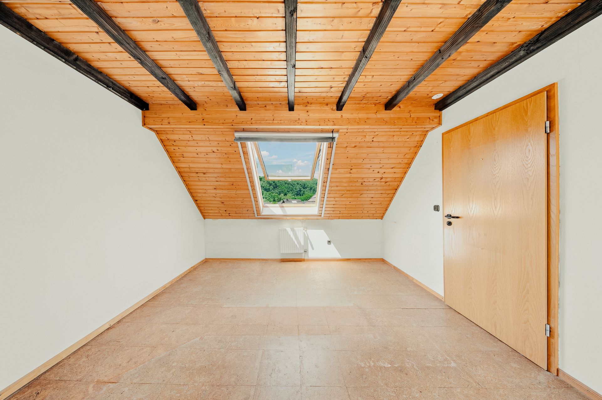 Leerer Dachraum mit Holzbalkendecke, großem Fenster und Holztür, Blick ins Grüne.