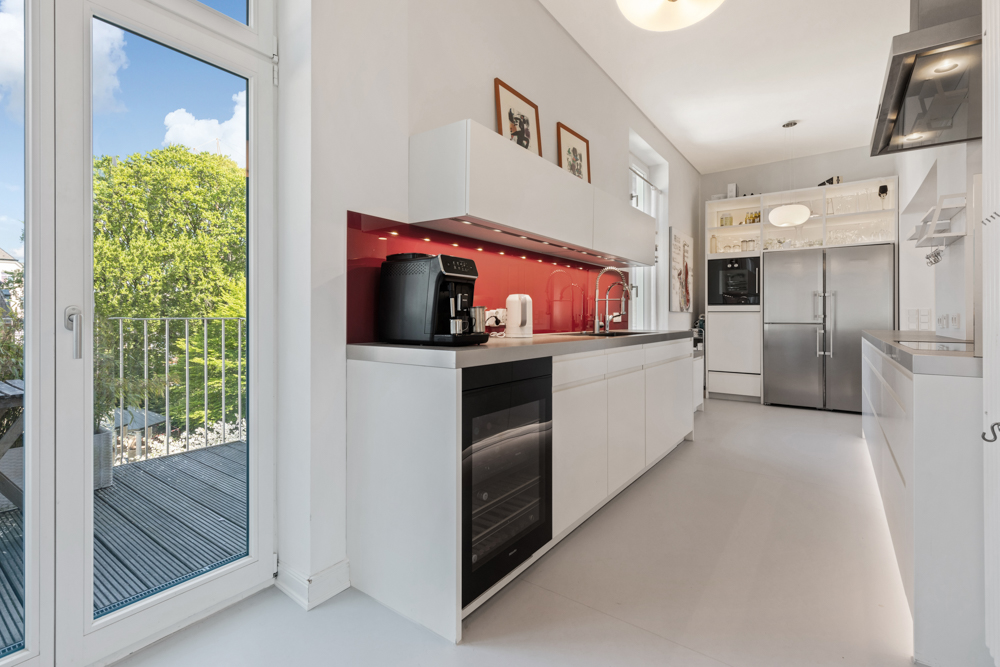 Moderne weiße Küche mit roter Rückwand, großem Fenster und Blick auf einen Balkon.