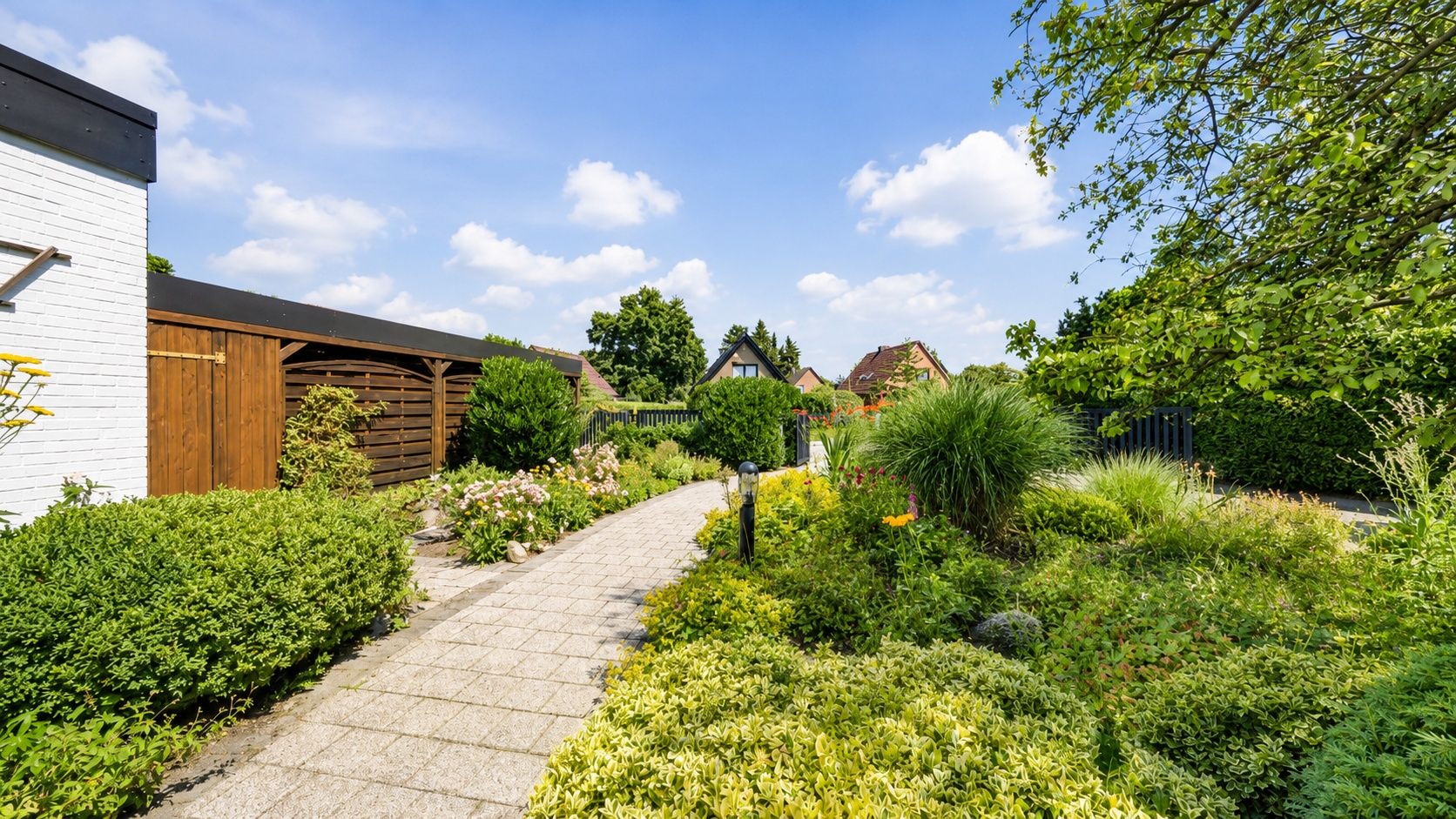 Gepflasterter Weg durch einen gepflegten Garten mit vielen Sträuchern und Blumen bei Sonnenschein.