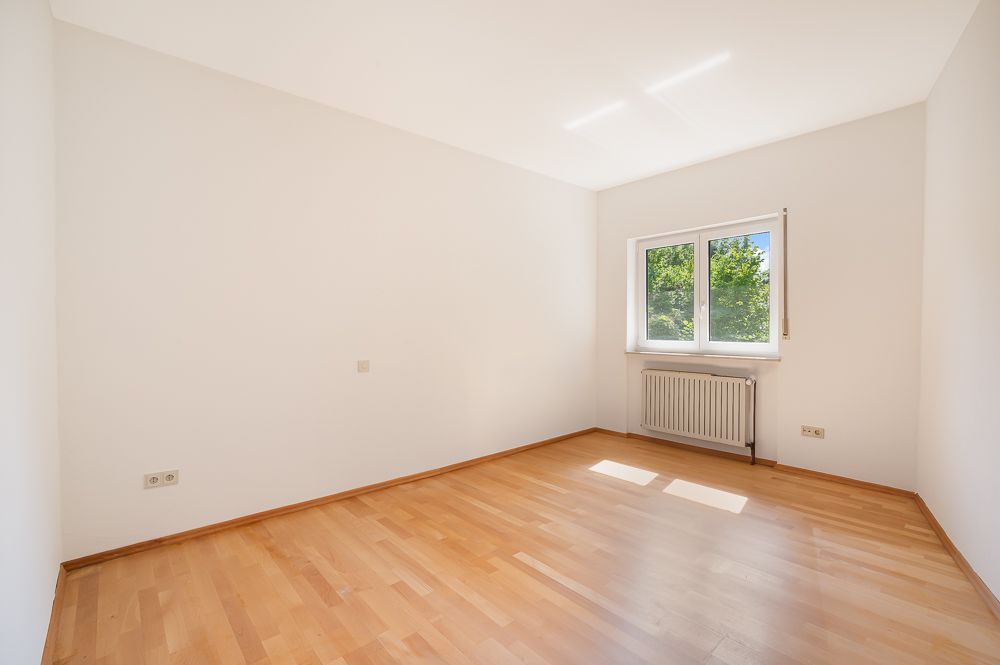 Leeres Zimmer mit Holzfußboden, weiß gestrichenen Wänden und einem Fenster mit Blick ins Grüne.