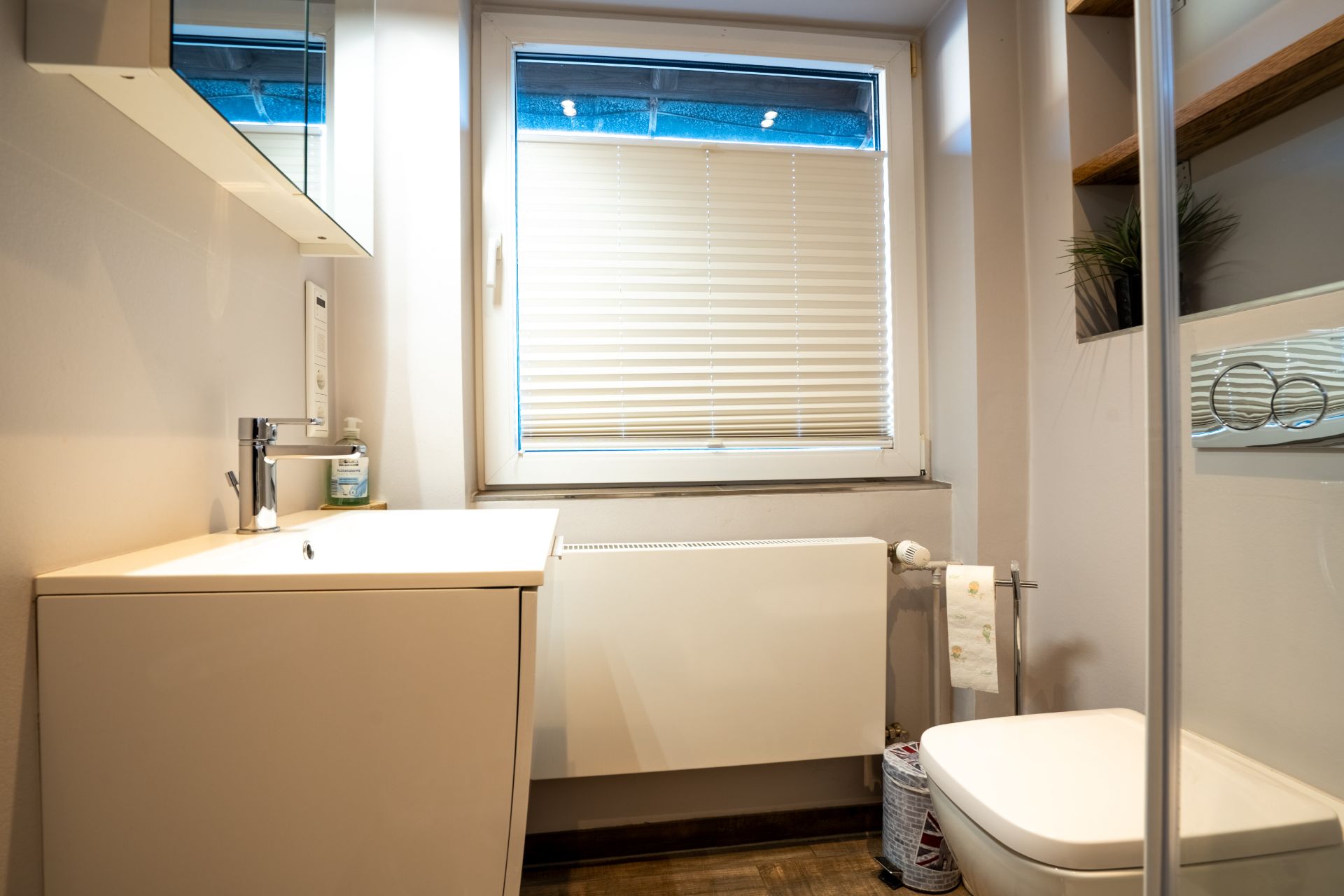Kleines Badezimmer mit Waschbecken, WC, Fenster mit Jalousie und Regal mit Pflanzen.