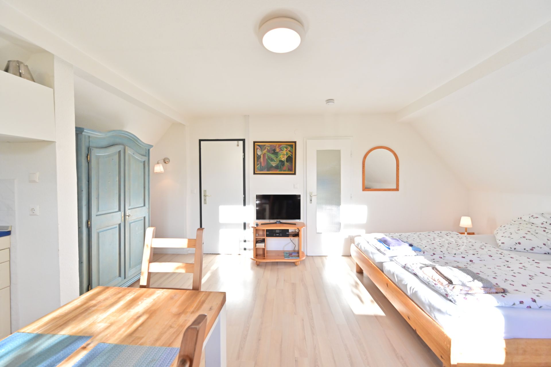 Helles Schlafzimmer mit Doppelbett, Holztisch, blauem Schrank und Fernseher an der Wand.