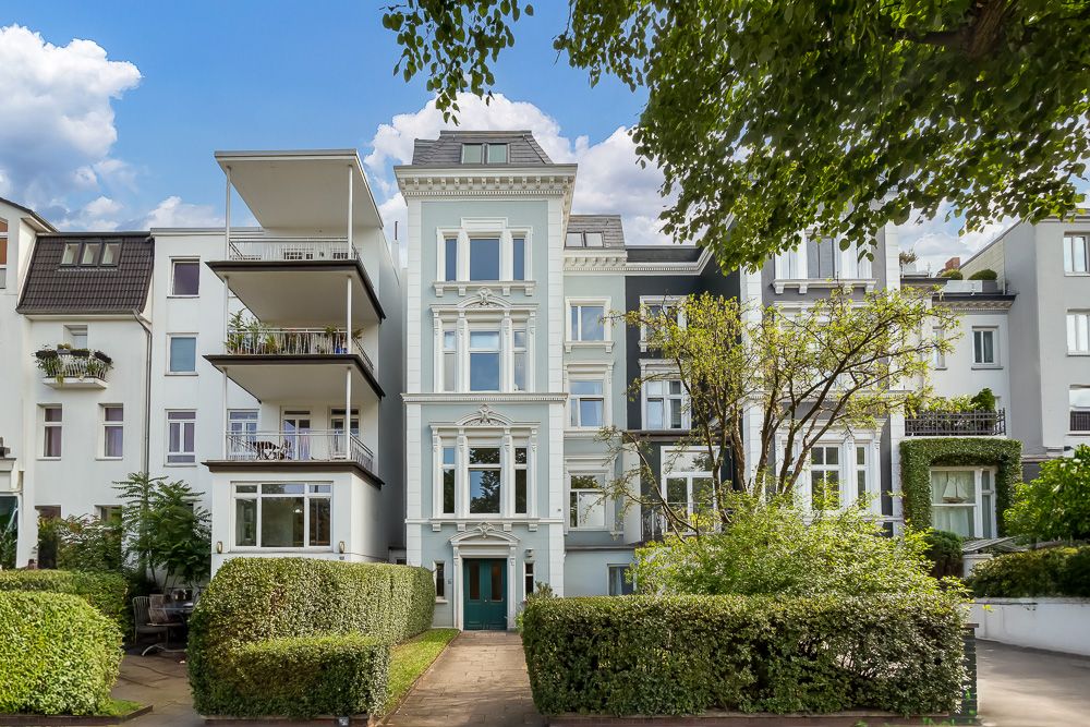 Mehrstöckiges Wohnhaus mit Balkonen, umgeben von Bäumen und gepflegten Hecken.