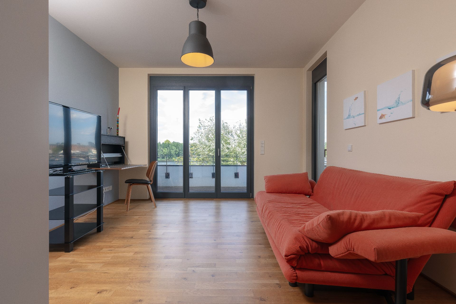 Wohnzimmer mit rotem Sofa, Fernseher, Schreibtisch und großen Fenstern zu einem Balkon.