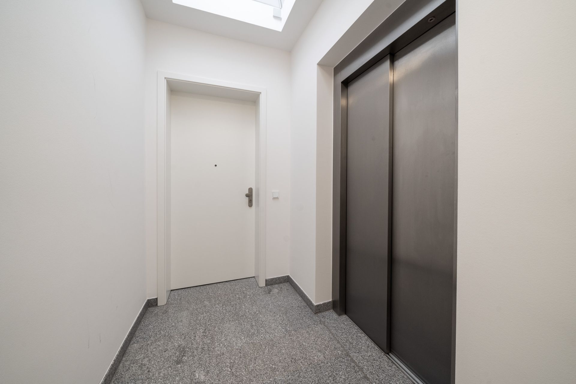 Flur mit weißer Tür und geschlossenem Aufzug, Tageslicht durch Oberlicht.