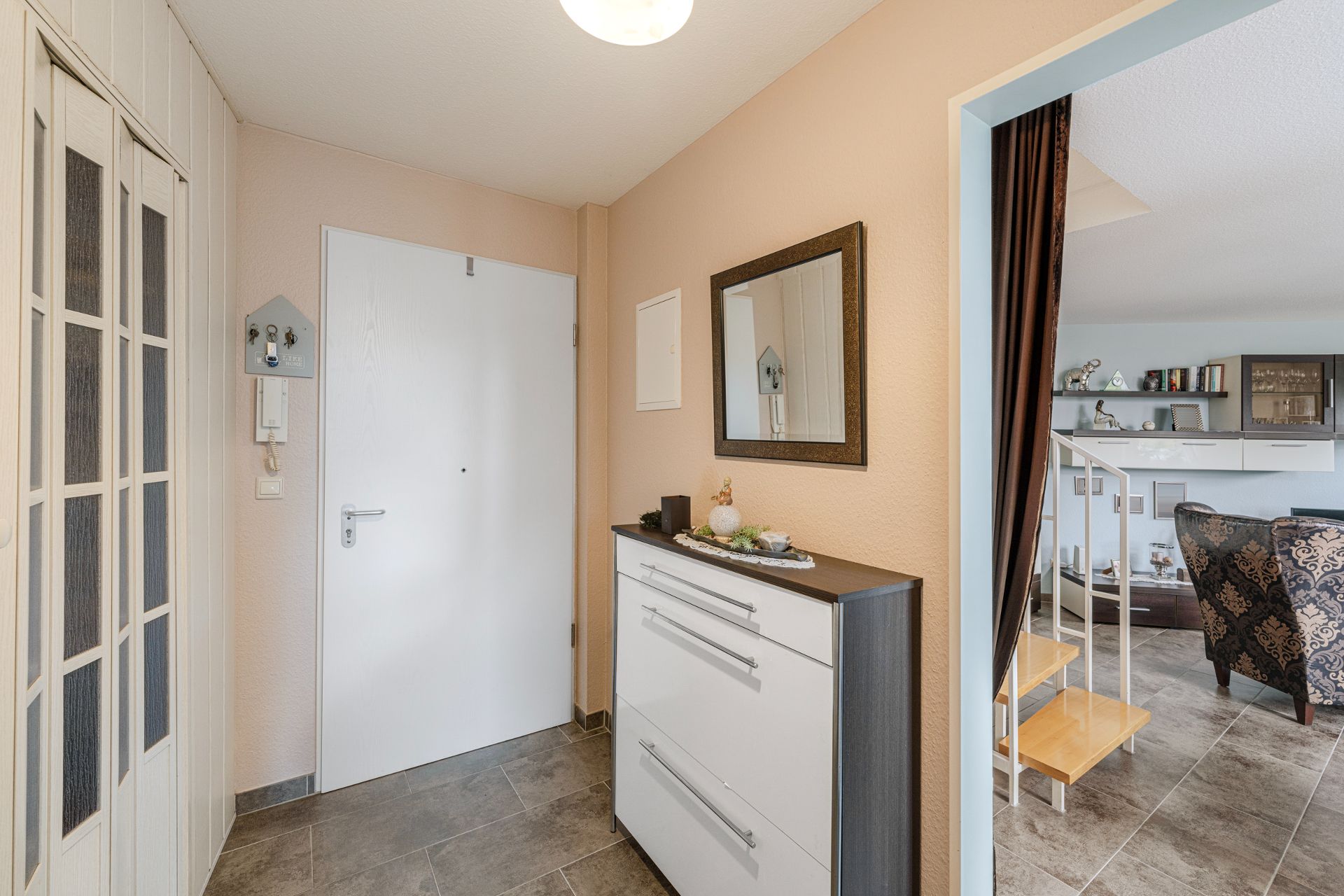 Heller Flur mit weißer Tür, Spiegel, Kommode und Blick ins Wohnzimmer mit Treppe.