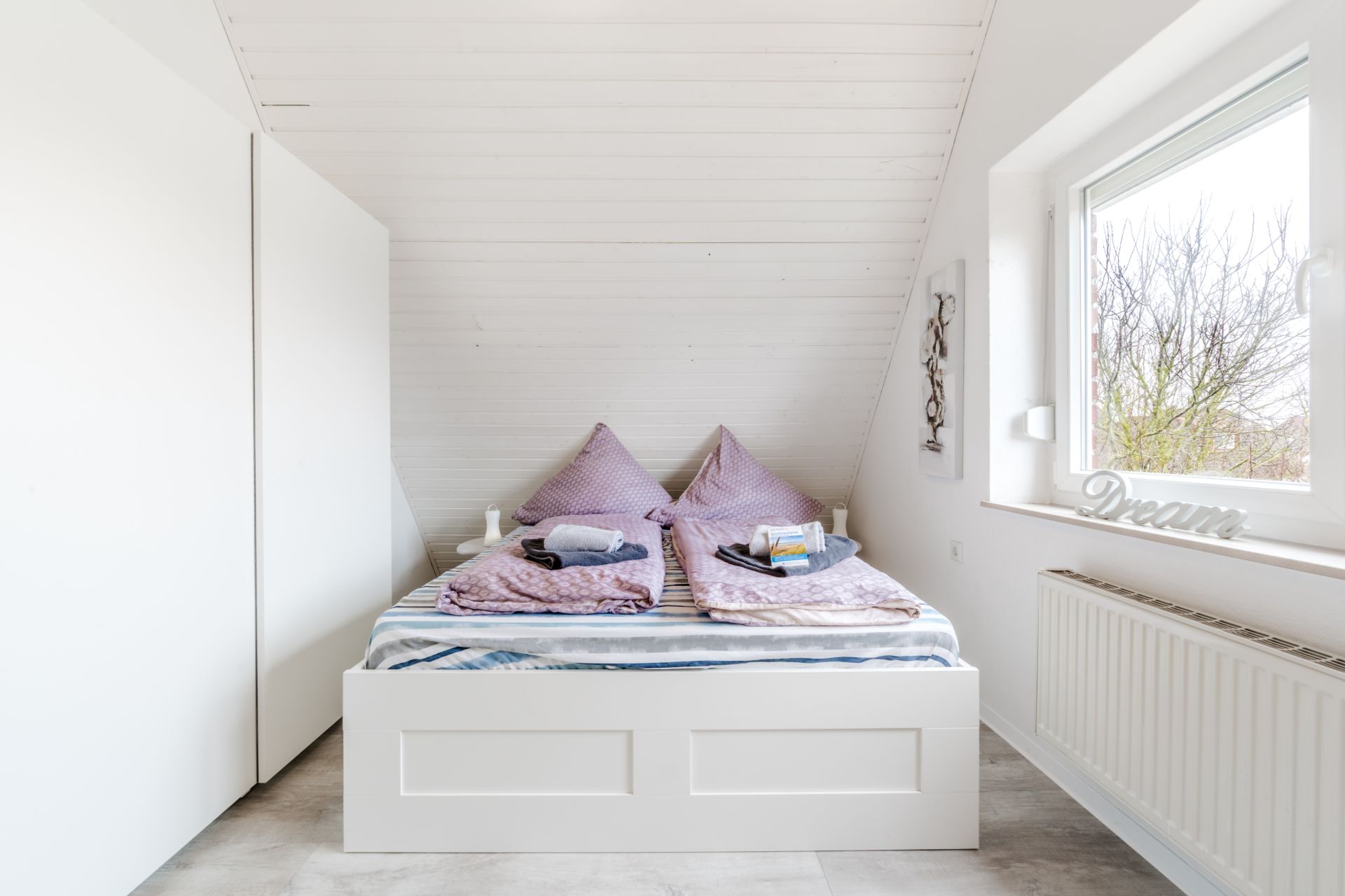 Helles Schlafzimmer mit Doppelbett, zwei lila Kissen, großem Fenster und weißem Kleiderschrank.