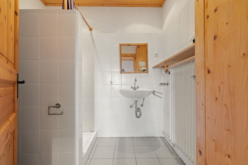 Kleines Badezimmer mit Dusche, Waschbecken und Spiegel, weiße Fliesen und Holzelemente.