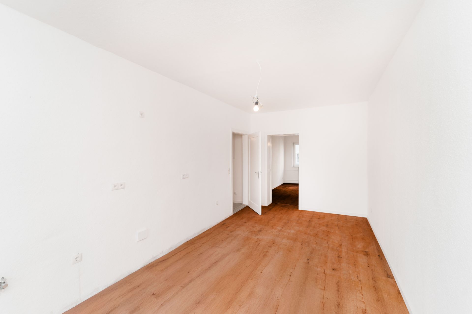 Leerer Raum mit weiß gestrichenen Wänden und Holzfußboden, eine offene Tür im Hintergrund.