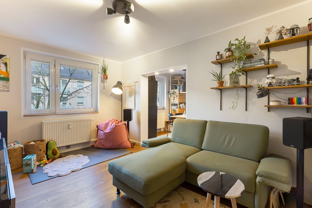 Wohnzimmer mit grünem Sofa, Wandregalen, Spielzeug und Blick in ein Esszimmer.