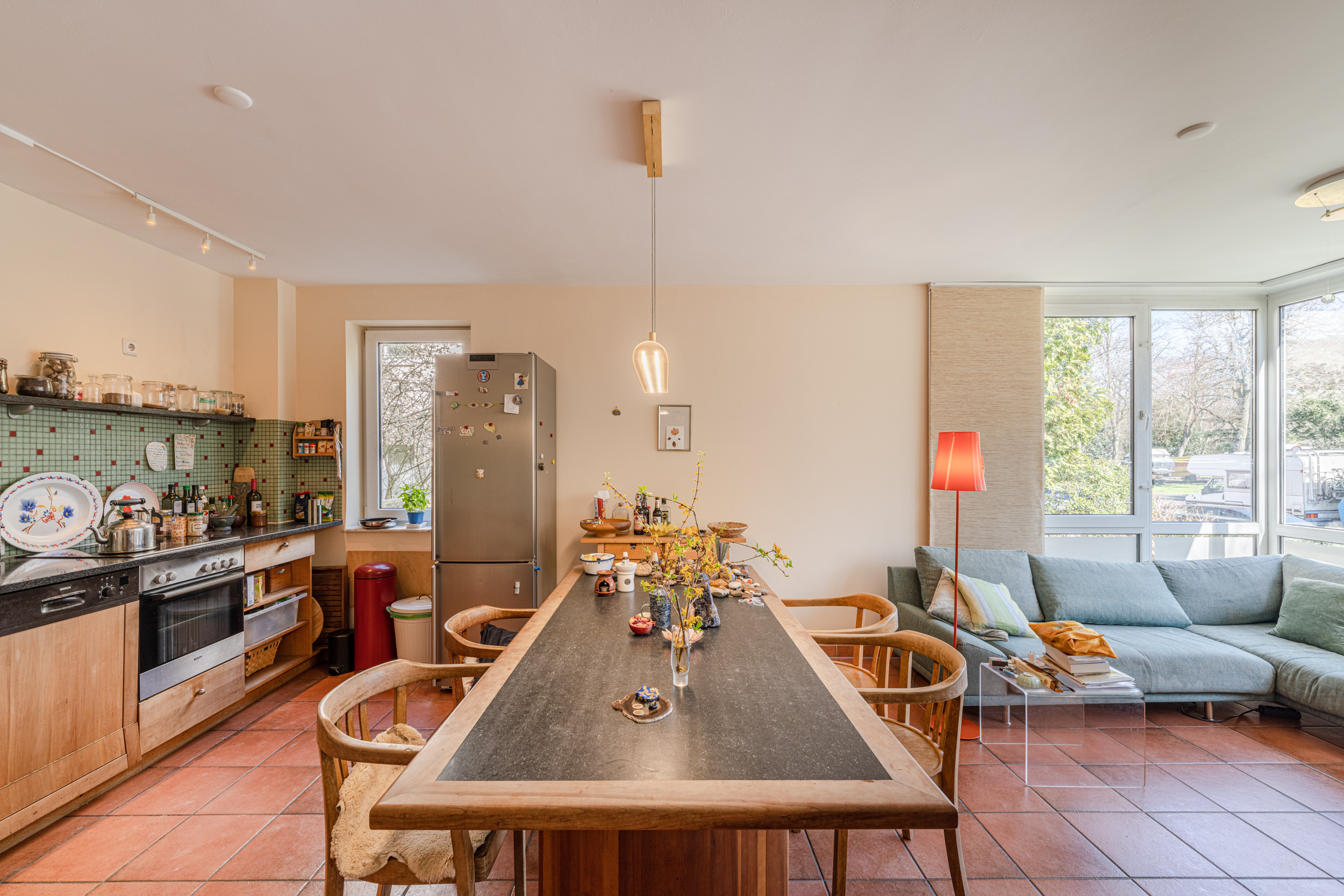 Helle Wohnküche mit Esstisch, Stühlen, Sofa und großen Fenstern mit Blick ins Grüne.
