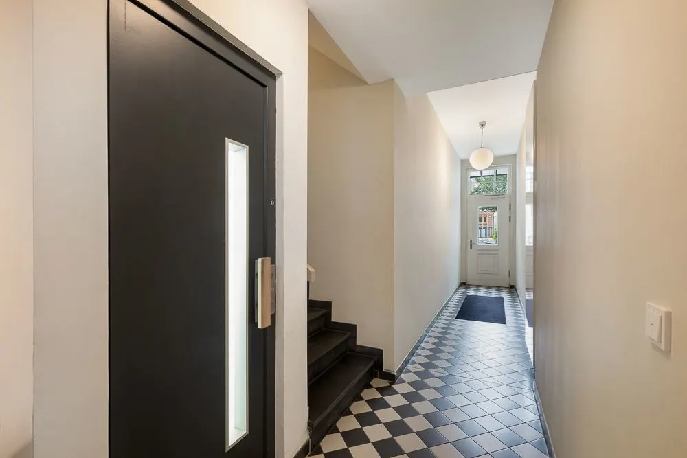 Heller Flur mit schwarz-weiß kariertem Boden, einer schwarzen Tür und einer Treppe.