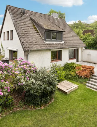 Weißes Einfamilienhaus mit Garten, bunten Blumen und Terrasse bei sonnigem Wetter.