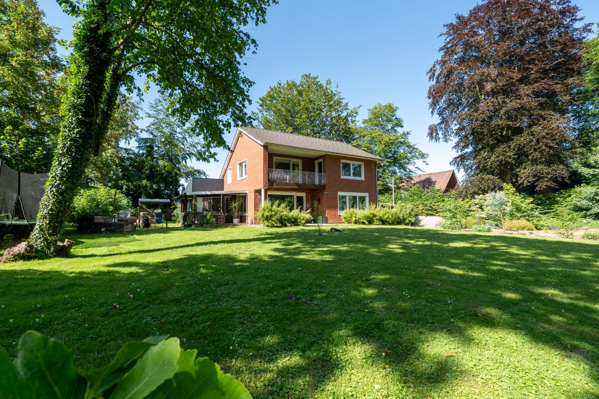Backsteinwohnhaus mit Balkon, großem Garten und vielen umliegenden Bäumen bei sonnigem Wetter.