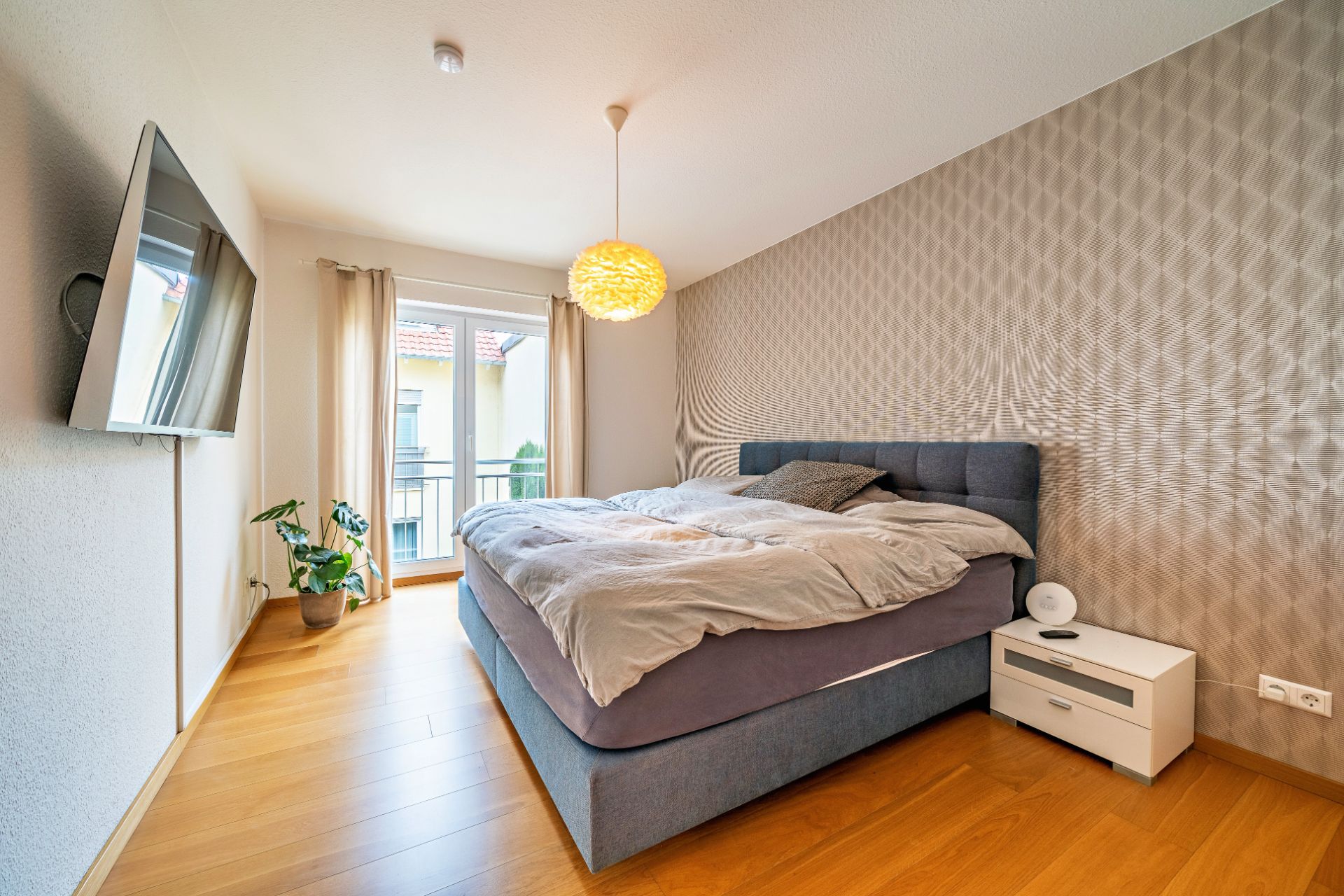 Modernes Schlafzimmer mit Doppelbett, Nachttisch, großem Fenster, Fernseher und Holzboden.