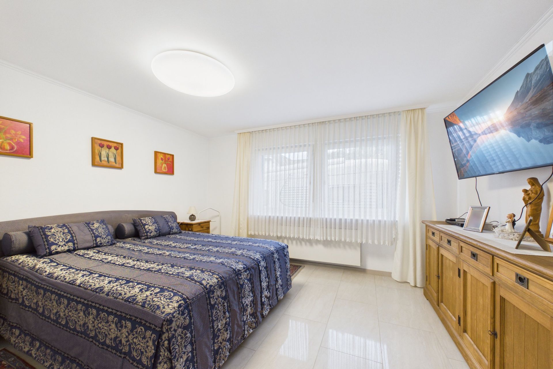 Schlafzimmer mit Doppelbett, Holzmöbeln, großem Fenster und Wandfernseher.
