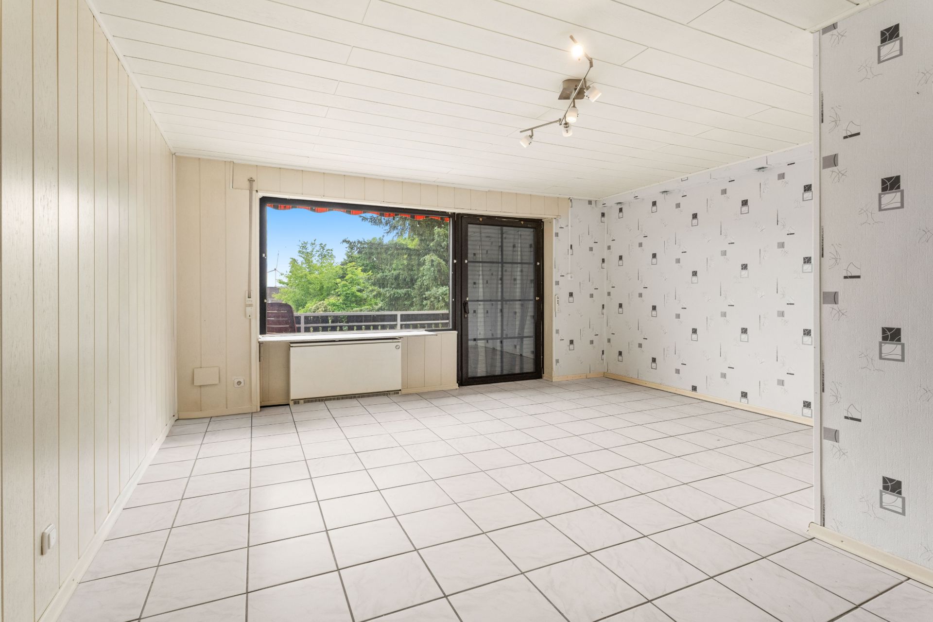 Leeres Wohnzimmer mit Fliesenboden, großen Fenstern und Glastür zum Balkon.