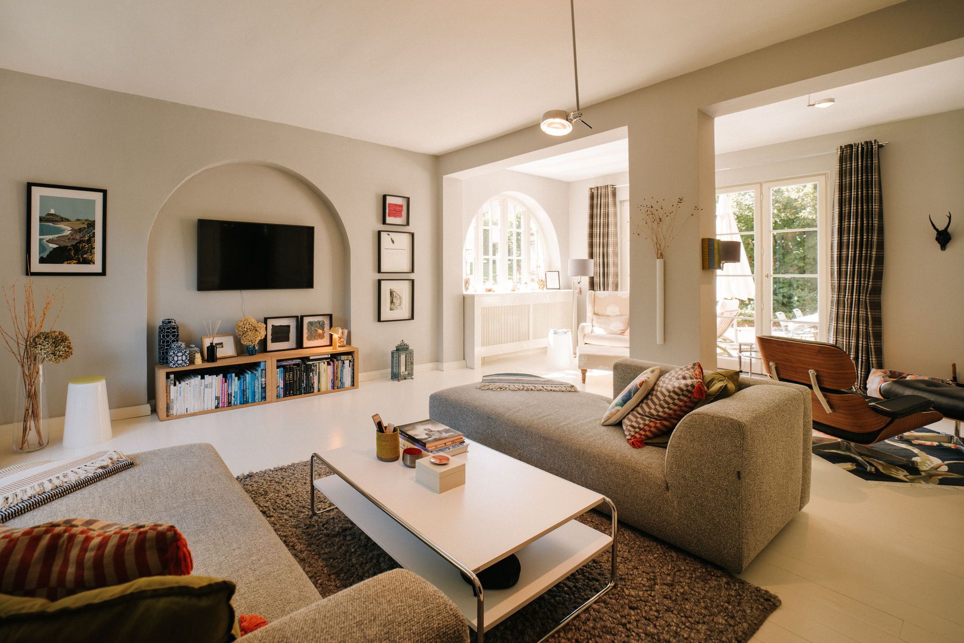 Helles Wohnzimmer mit grauen Sofas, Couchtisch, TV an der Wand und großen Fenstern.