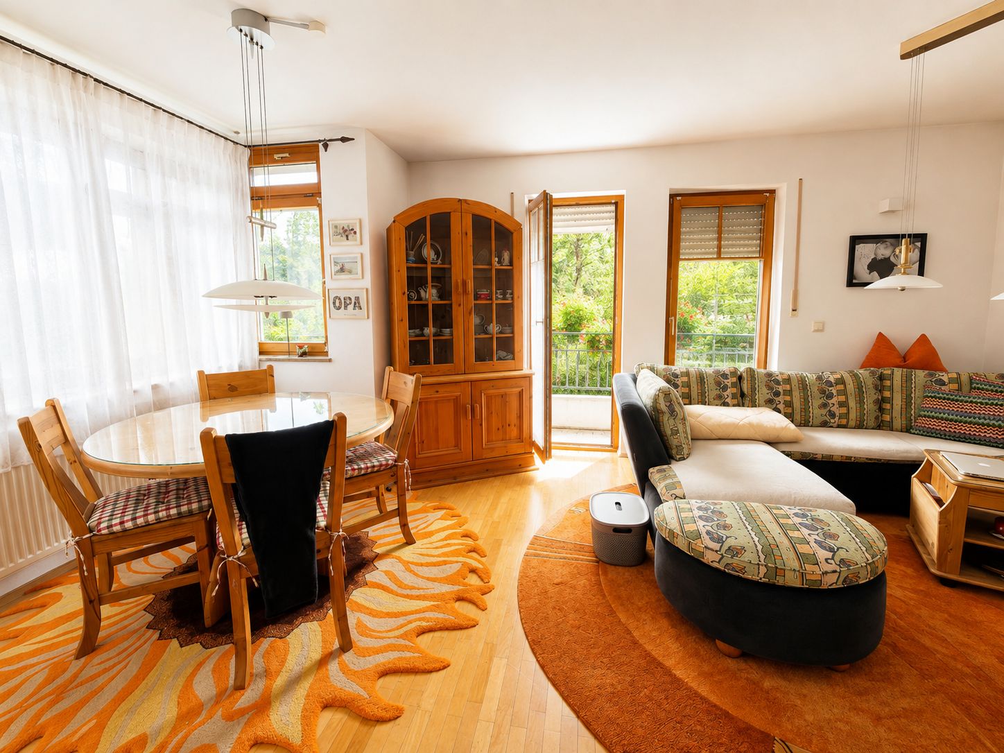 Wohnzimmer mit runder Couch, Esstisch, Holzschrank und Zugang zum Balkon, helle Fenster.