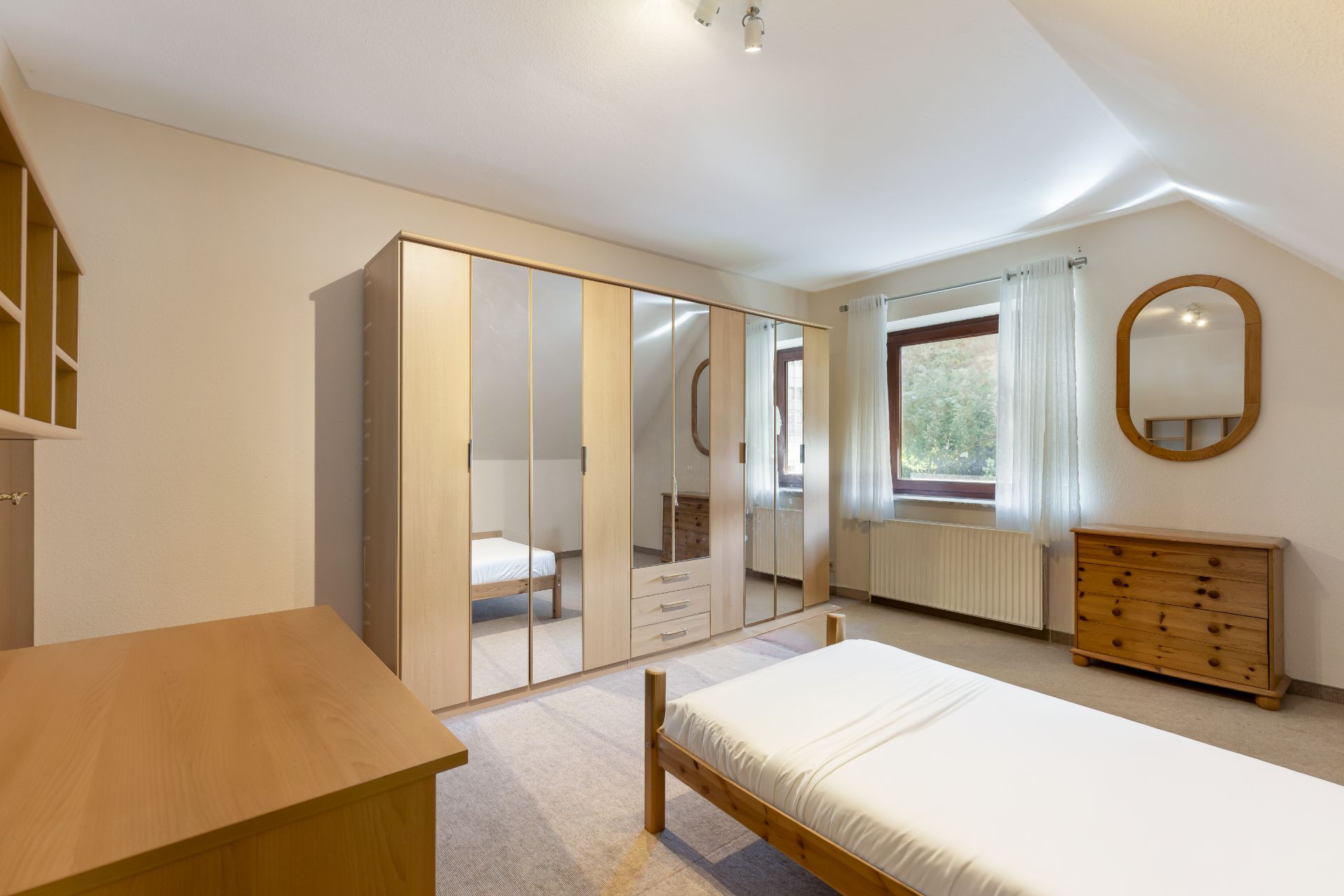 Helles Schlafzimmer mit Einzelbett, großem Spiegelschrank, Schreibtisch und Fenster mit Vorhängen.