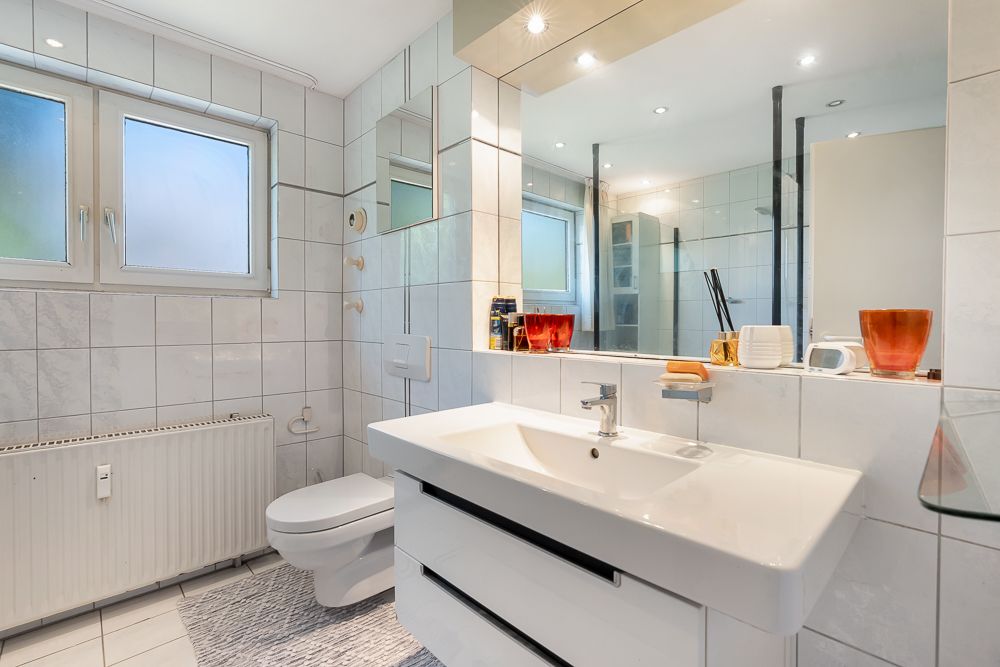 Helles Badezimmer mit großem Spiegel, Waschbecken, WC, zwei Fenstern und weißen Fliesen.