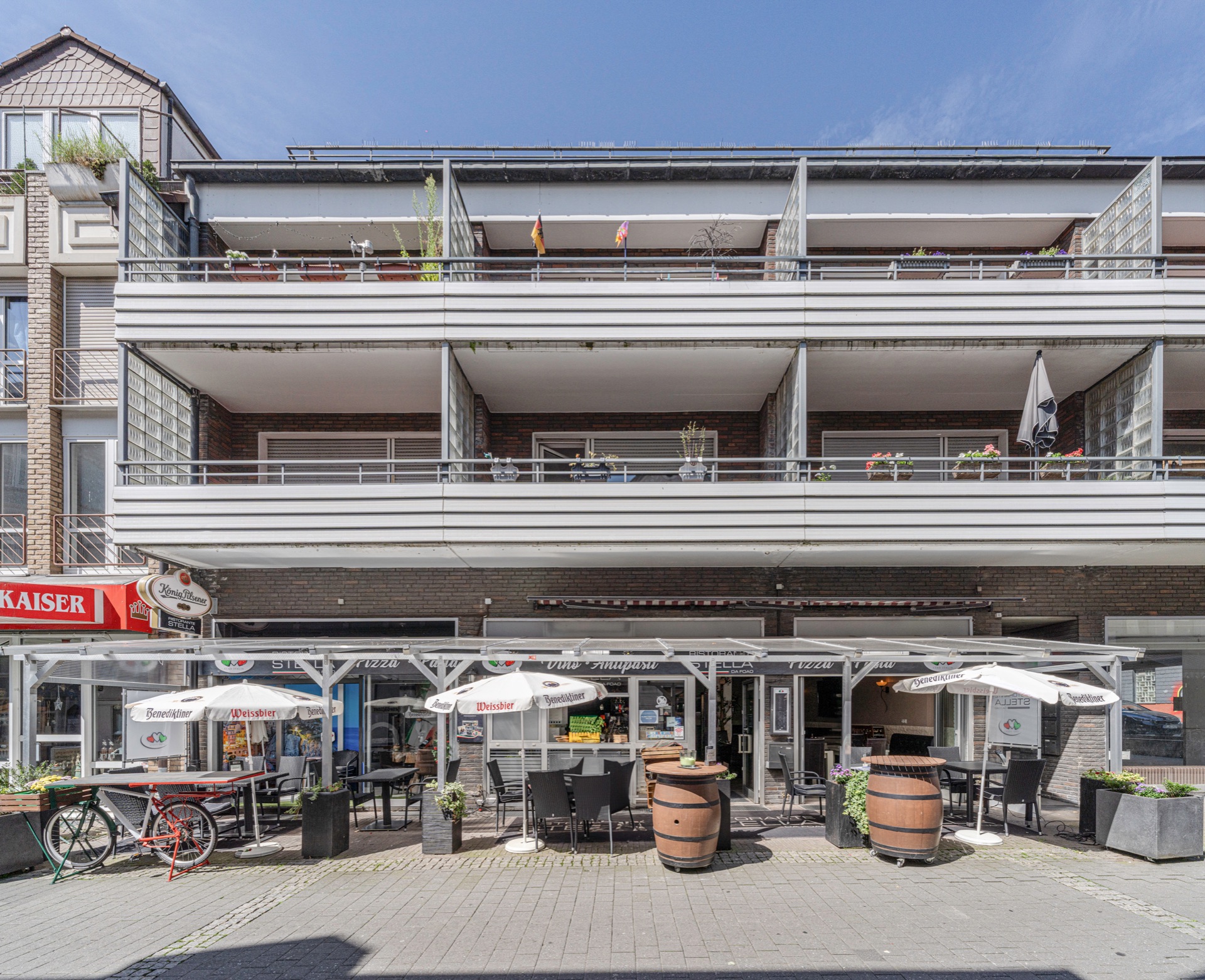 Mehrstöckiges Wohnhaus mit Balkonen und Straßencafé im Erdgeschoss, Tische und Sonnenschirme draußen.