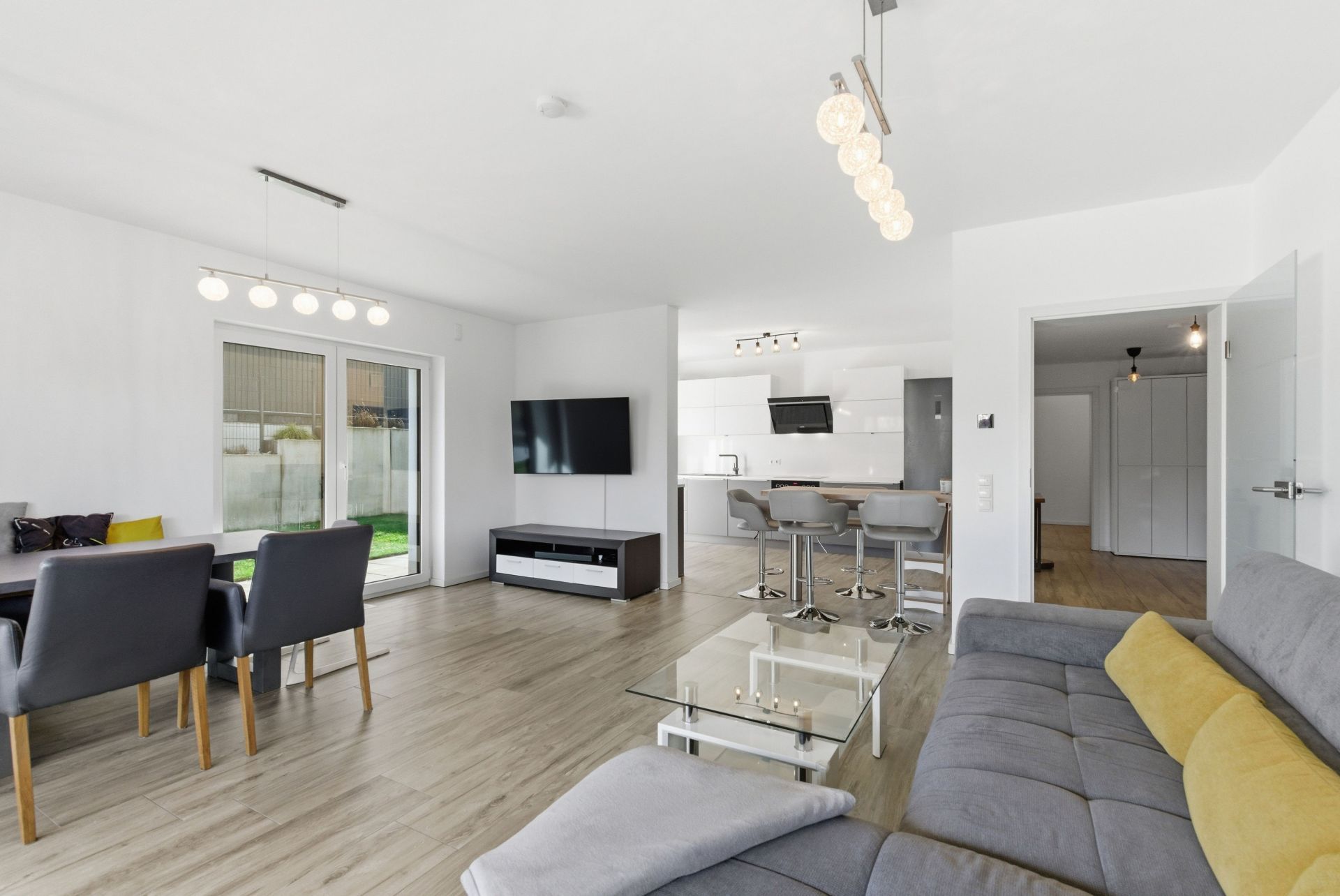 Modernes Wohnzimmer mit grauem Sofa, Glastisch, Esstisch, TV und offener Küche.