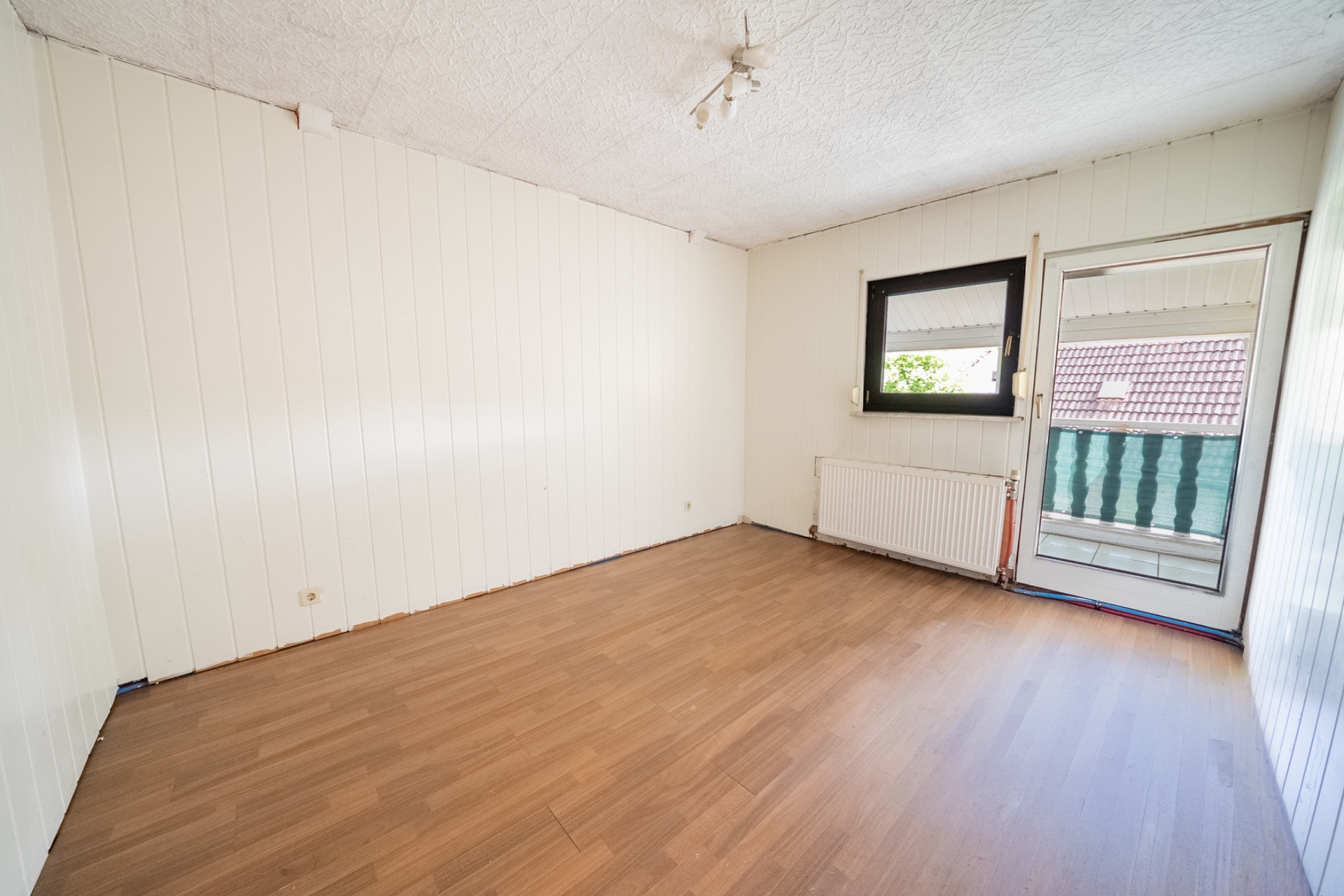 Leeres Zimmer mit Holzfußboden, weißer Wand und Fenster sowie Tür zu einem Balkon.