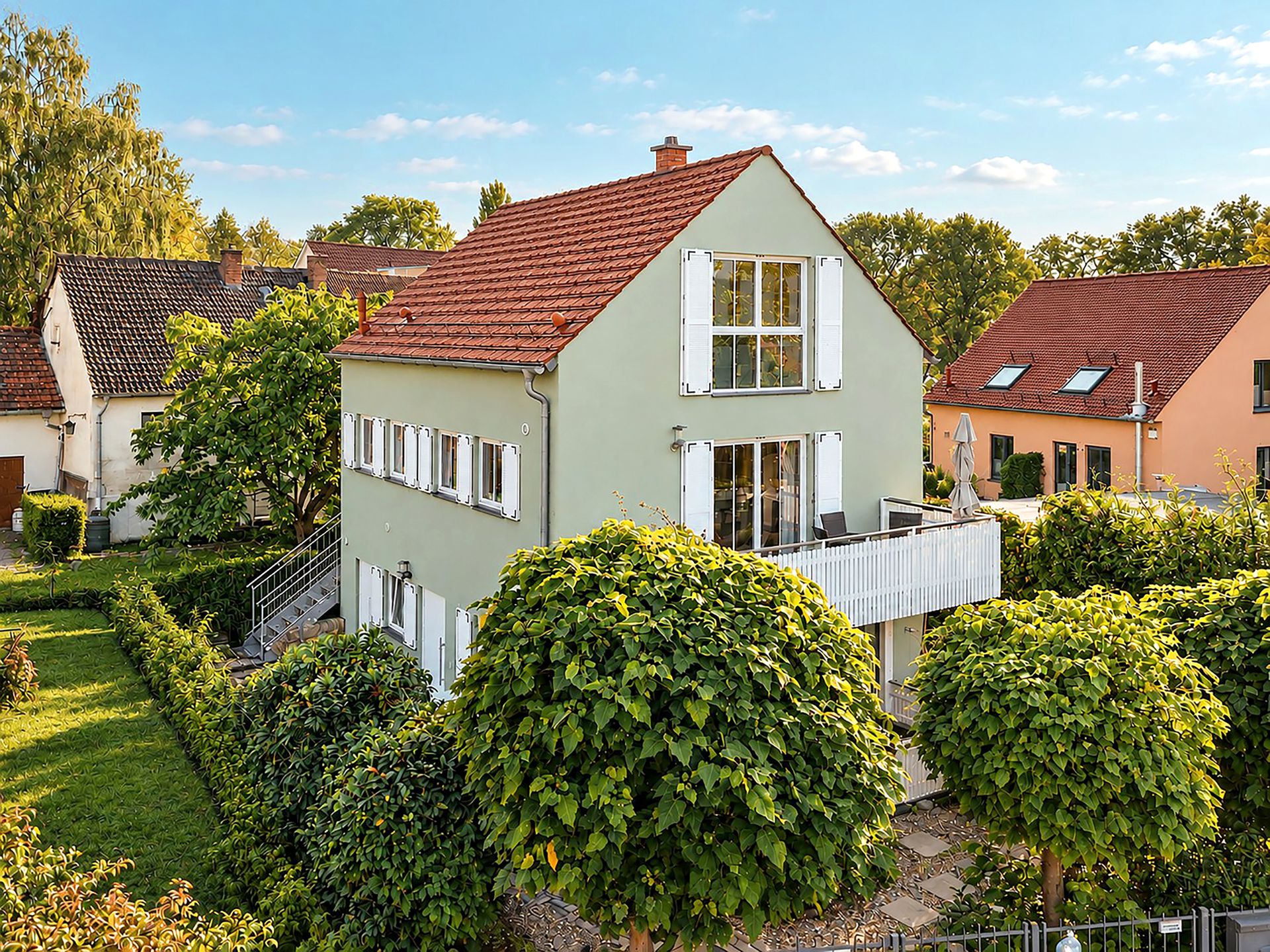 Zweistöckiges Wohnhaus mit Balkon, Garten und Bäumen im Vordergrund bei sonnigem Wetter.