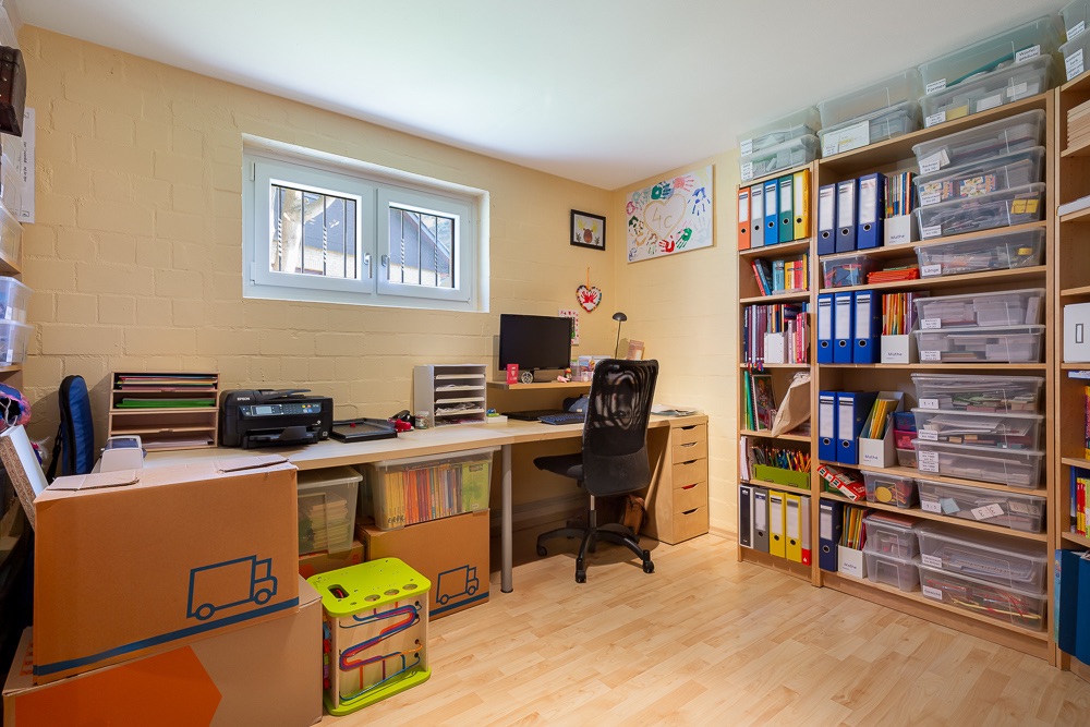 Arbeitszimmer mit Schreibtisch, Regal voller Ordner, Umzugskartons und Computer.
